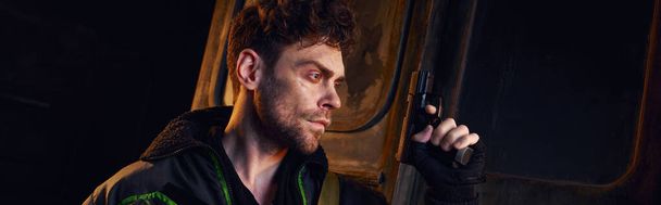 Ongeschoren man in versleten jasje staand met pistool in donkere ondergrondse, post-apocalyptische concept, spandoek - Foto, afbeelding
