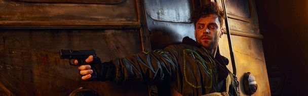 небритый игровой персонаж в изношенной одежде, нацеленный с пистолетом в опасном постапокалиптическом метро, баннер - Фото, изображение