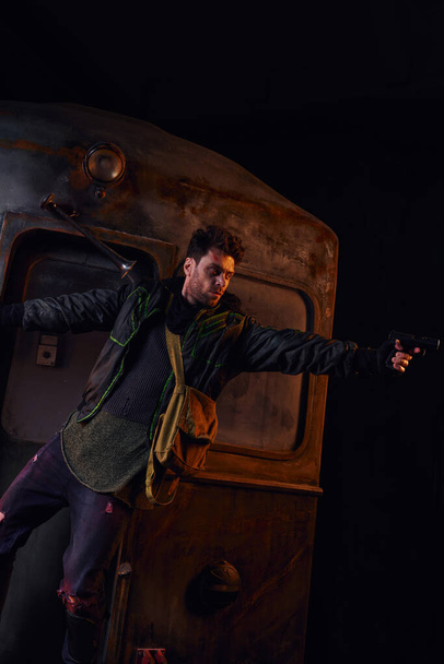 Unrasierter Mann in verschlissenem Outfit zielt mit Spaß auf rostigen Waggon in der Dunkelheit der U-Bahn nach der Katastrophe - Foto, Bild