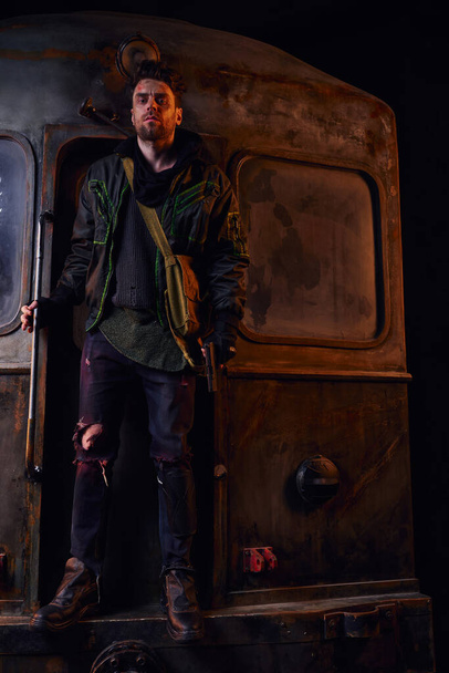 hombre en traje desgastado sosteniendo la pistola y mirando a la cámara cerca del vagón oxidado del metro, la vida después del desastre - Foto, Imagen