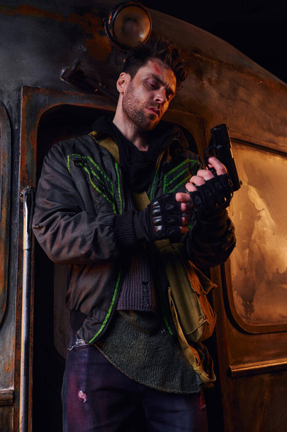 poważny mężczyzna w noszonej kurtce i rękawiczki bez palców trzymając pistolet w pobliżu zardzewiałego powozu w opuszczonym metrze - Zdjęcie, obraz