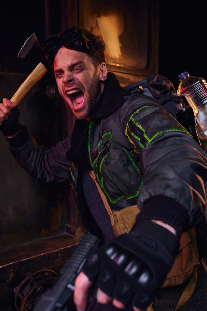 hombre herido sin afeitar con hacha y pistola gritando y defendiendo en el oscuro metro post-apocalíptico - Foto, imagen