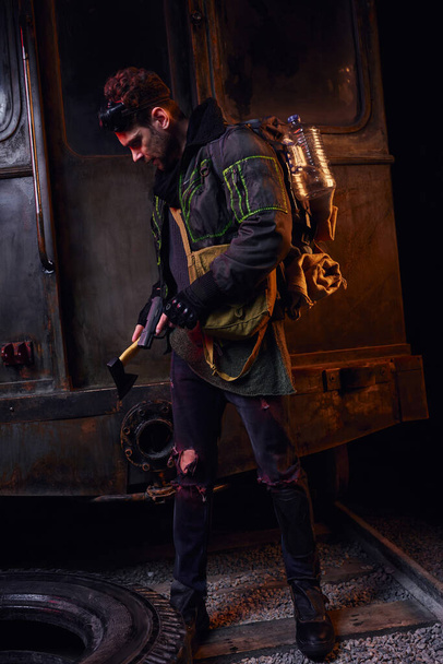 Mann mit Waffe und Axt bewaffnet steht mit Rucksack und Wasserflasche in dunkler verlassener U-Bahn - Foto, Bild