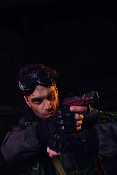 концентрована людина з військовими окулярами, спрямована з пістолетом у темряві пост-апокаліптичного підпілля - Фото, зображення