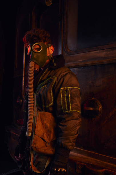 человек в грязной куртке и противогазе возле ржавого вагона метро в заброшенном подземном туннеле - Фото, изображение
