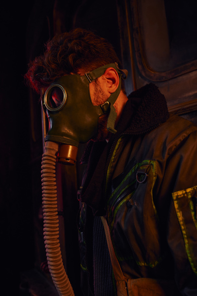 hombre con máscara y chaqueta desgastada en luz roja de túnel subterráneo oscuro, supervivencia post-apocalíptica - Foto, imagen