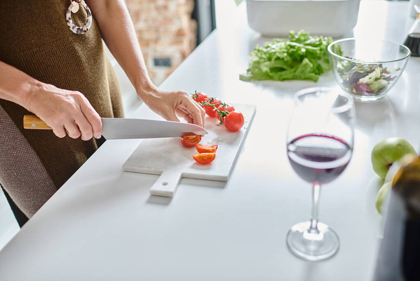 частичный снимок женщины, режущей помидоры черри возле бокала красного вина и салата в прозрачной миске - Фото, изображение