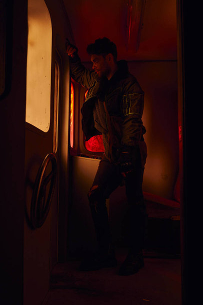 hombre con arma mirando a través de la ventana del carruaje abandonado en el metro post-desastre con luz roja - Foto, Imagen