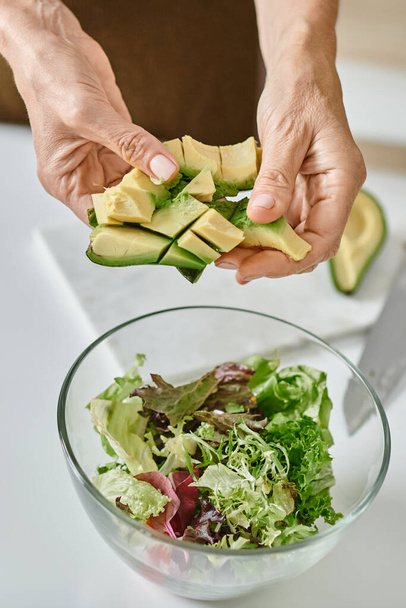 жінка тримає нарізаний стиглий авокадо біля салату в скляній мисці, крупним планом знімок жіночих рук - Фото, зображення