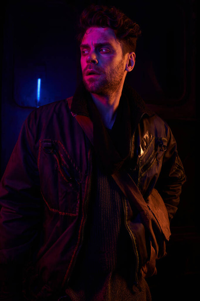 tense man in black jacket looking away in darkness of abandoned underground tunnel with neon light - Photo, Image