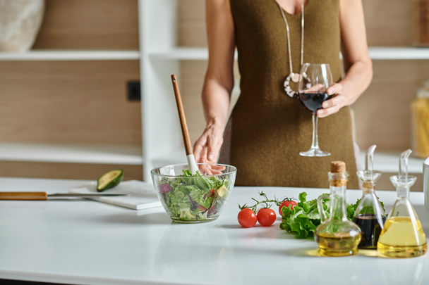 Kırpılmış kadın elinde bir kadeh kırmızı şarap tutuyor ve şeffaf kasede taze yapılmış salatanın yanında duruyor. - Fotoğraf, Görsel