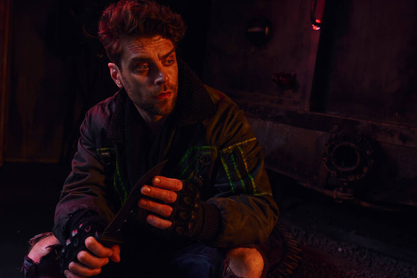 στοχαστικός άνθρωπος με γάντια χωρίς δάχτυλα κάθεται με στρατιωτικό μαχαίρι σε σκούρο υπόγειο με κόκκινο φως - Φωτογραφία, εικόνα