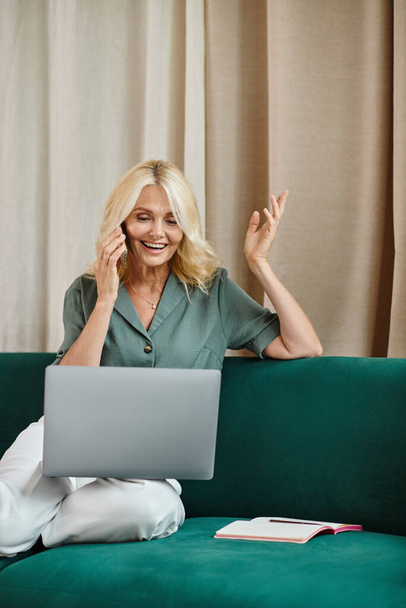 cheerful middle aged woman with blonde hair talking on smartphone and using laptop on sofa - Fotó, kép