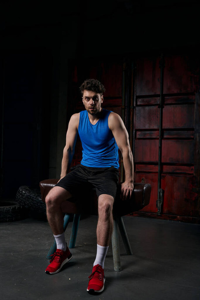 уверенный в себе мужчина в спортивной одежде, смотрящий в камеру и сидящий на спортивной скамейке ночью, полной длины - Фото, изображение
