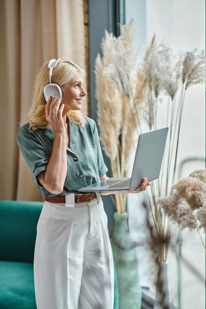 θετική μεσήλικη γυναίκα με ασύρματα ακουστικά που ακούει μουσική και χρησιμοποιεί φορητό υπολογιστή στο σπίτι - Φωτογραφία, εικόνα