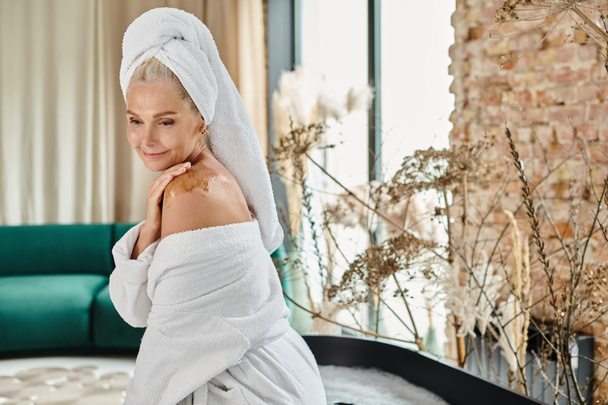 jolie femme d'âge moyen avec serviette blanche sur la tête et peignoir en utilisant gommage du corps sur l'épaule - Photo, image