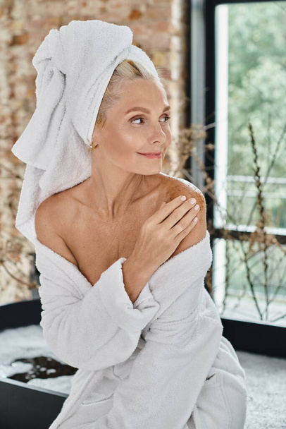 mujer de mediana edad complacida con toalla blanca en la cabeza y albornoz aplicando exfoliación corporal en el hombro - Foto, Imagen