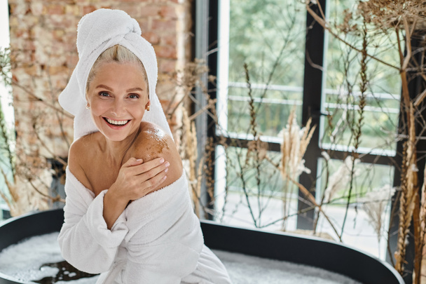 mulher de meia-idade alegre com toalha branca na cabeça e roupão de banho aplicando esfoliação corporal perto da banheira - Foto, Imagem