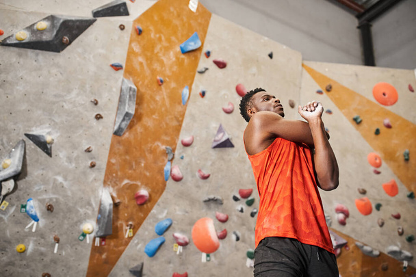 athletic african american man in orange shirt warming up with rock climbing wall backdrop, sport - Photo, Image