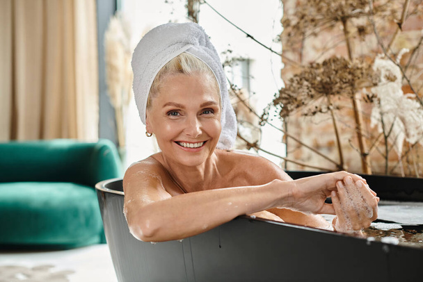 mujer de mediana edad feliz con toalla blanca en la cabeza tomando baño en apartamento moderno, relajación - Foto, imagen