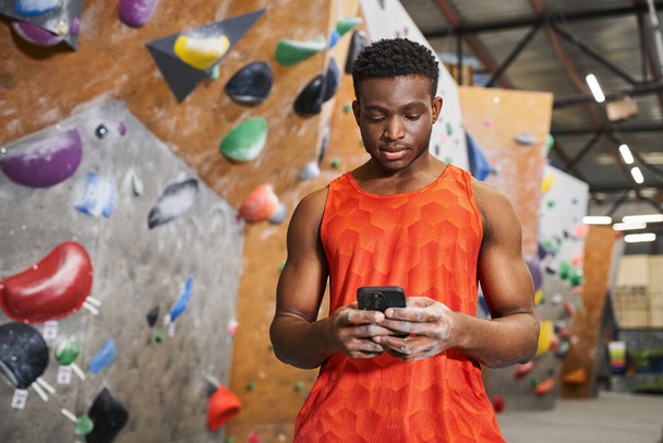 koncentrált afro-amerikai férfi narancssárga ingben nézi a mobiltelefonját, sziklamászás - Fotó, kép