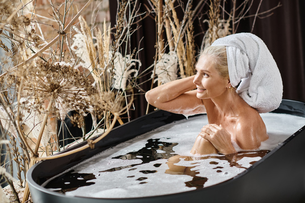 расслабленной женщины среднего возраста с белым полотенцем на голове принимая ванну в современной квартире, красота рутины - Фото, изображение