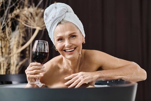 alegre mujer de mediana edad con toalla en la cabeza sosteniendo un vaso de vino tinto mientras toma un baño en casa - Foto, imagen