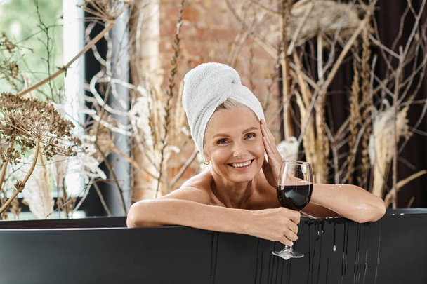 rento ja onnellinen keski-ikäinen nainen pyyhe pään tilalla lasi punaviiniä kylvyssä - Valokuva, kuva