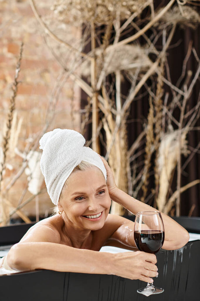 мрійлива жінка середнього віку з рушником на голові тримає келих червоного вина під час прийняття ванни - Фото, зображення