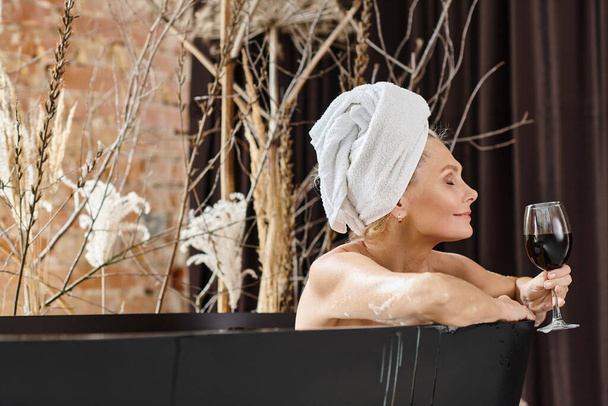 mujer de mediana edad relajada y alegre con toalla en la cabeza sosteniendo un vaso de vino tinto y tomando un baño - Foto, Imagen