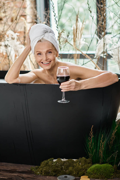 mujer radiante de mediana edad con toalla blanca en la cabeza sosteniendo vaso de vino tinto y tomando baño - Foto, Imagen
