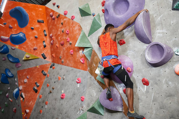 sportieve Afro-Amerikaanse man in oranje shirt met alpine harnas klimmen muur, sport concept - Foto, afbeelding