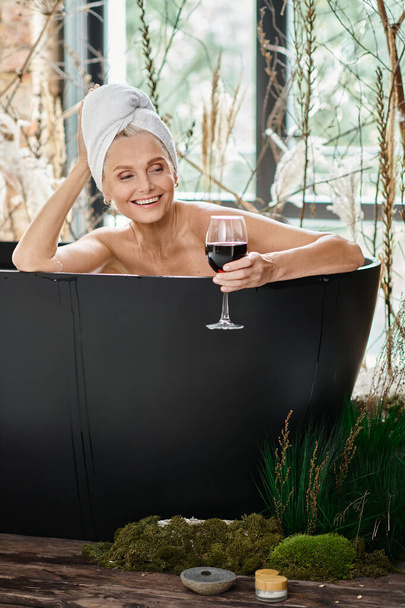 Kafasında beyaz havluyla gülümseyen orta yaşlı bir kadın elinde bir bardak kırmızı şarap ve banyo yapıyor. - Fotoğraf, Görsel