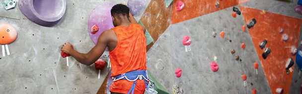 sporty african american man in orange shirt climbing up rock wall with alpine harness, banner - Photo, Image
