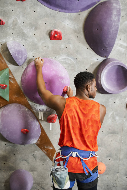colpo verticale di uomo muscoloso afroamericano in camicia arancione arrampicata su parete rocciosa, bouldering - Foto, immagini
