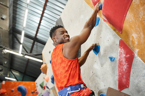cheerful african american man smiling happily at camera while climbing up rock wall, bouldering - Photo, Image