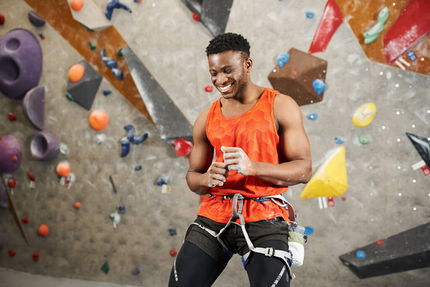 alegre africano americano hombre en naranja camisa riendo sinceramente posando junto a roca escalada pared - Foto, Imagen