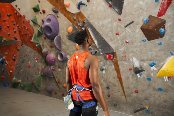 uomo afroamericano sportivo in camicia arancione con imbracatura alpina pronta a scalare parete rocciosa, vista posteriore - Foto, immagini