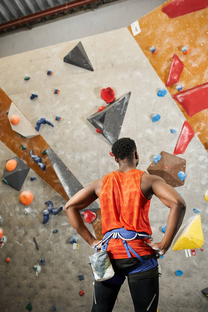 назад вид спортивного африканского американца в оранжевой рубашке с альпийской упряжкой с руками акимбо - Фото, изображение