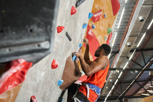 handsome african american man in orange shirt with alpine harness climbing up bouldering wall - Photo, Image