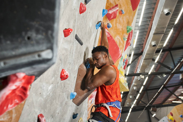 handsome young african american male model with alpine harness climbing up bouldering wall - Photo, Image