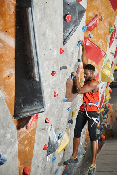 vertical shot of sporty african american man in orange shirt hanging on his fingers on climbing wall - Photo, Image