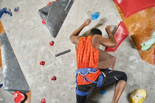 athletic young african american man with alpine harness ascending up bouldering wall, back view - Photo, Image