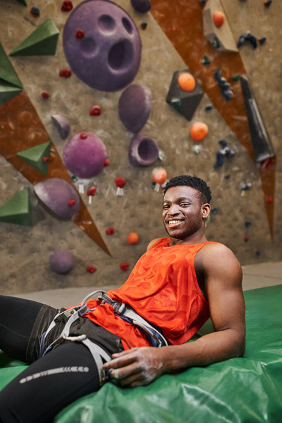 plano vertical de alegre afroamericano relajante en almohadilla de bouldering y sonriendo alegremente a la cámara - Foto, imagen