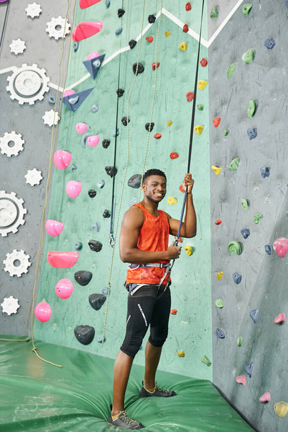 cheerful african american man standing on crash pad using climbing rope and smiling at camera - Photo, Image