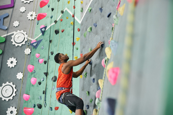 young african american man climbing up bouldering wall with safety rope and alpine harness - Photo, Image