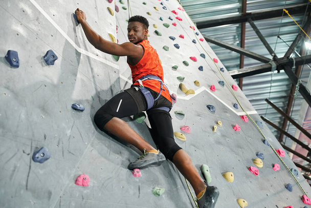forte uomo afroamericano in camicia arancione arrampicata su parete con corda di sicurezza e guardando giù - Foto, immagini