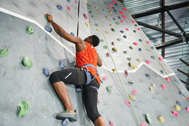 gespierde Afro-Amerikaanse man met alpine harnas en veiligheidstouw klimmen bouldering muur - Foto, afbeelding