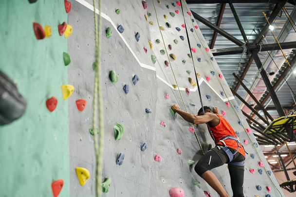 deportivo joven africano americano hombre en naranja camisa ascendiendo hasta bouldering pared con cuerda de seguridad - Foto, Imagen