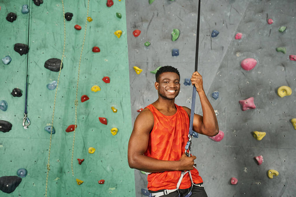 cheerful sporty african american man in orange shirt smiling joyfully at camera, bouldering concept - Photo, Image
